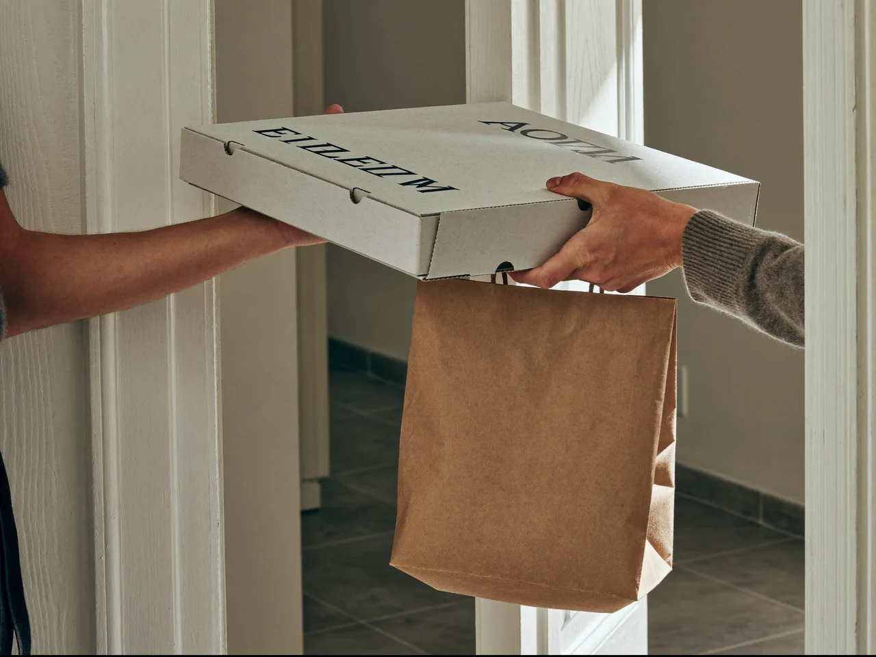 Übergabe von Pizzakarton und Papiertüte an einer Wohnungstür am Morgen